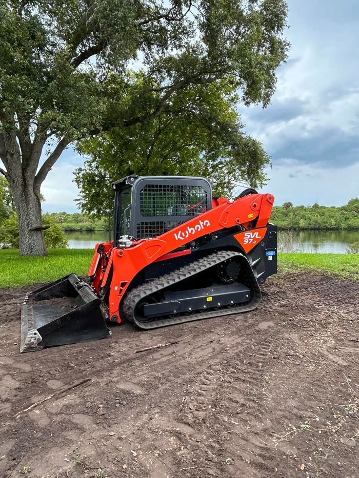 A red and black skid steer loader is parked in a dirt lot next to a tree.