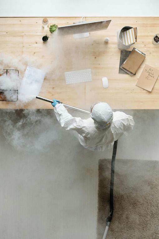 Une personne en combinaison de protection désinfecte un bureau et une moquette encombrés avec une substance semblable à un brouillard.
