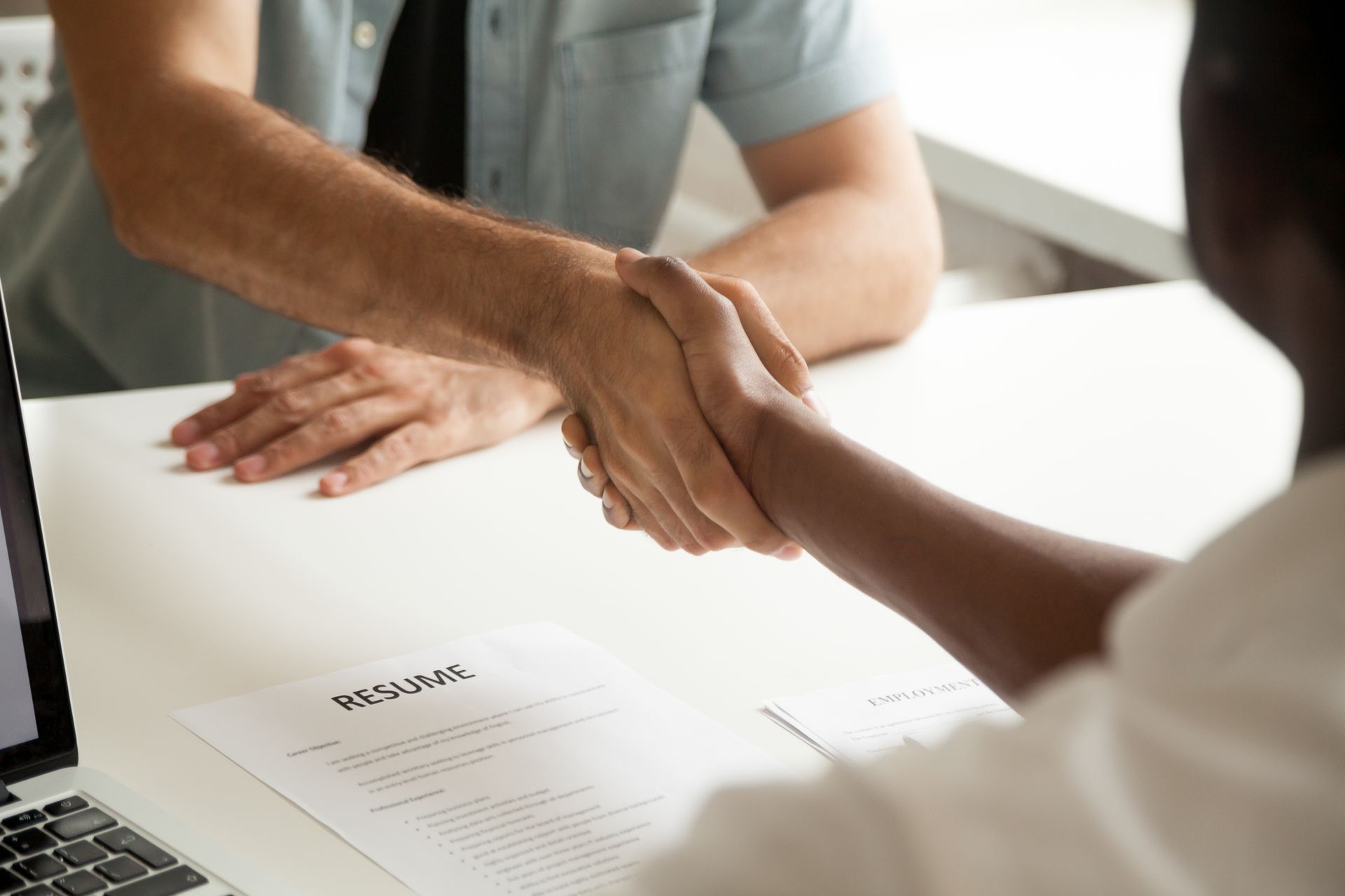 Two People Shaking Hands Across a White Desk With a Resume and Laptop — Search Partners in Melbourne, VIC