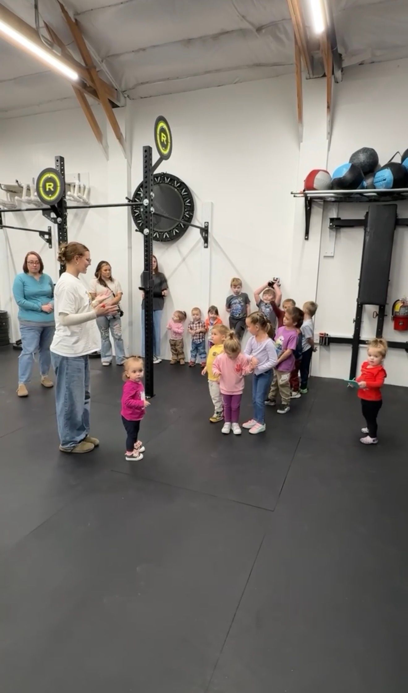 Children and adults gathered in Everyday Strength, a gym in Wenatchee, WA around a coach, with weights and fans on the wall.