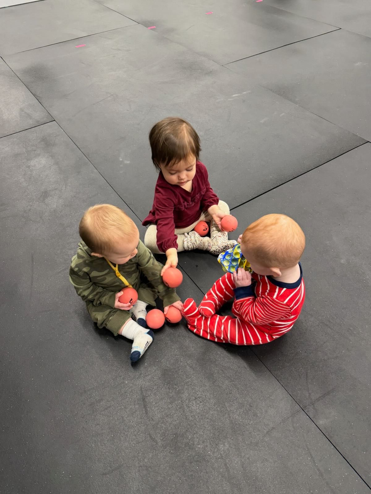 Three small children sit in a circle on dark playground tiles, playing with small balls and stuffed toys at Everyday Strength, a gym in Wenatchee, WA