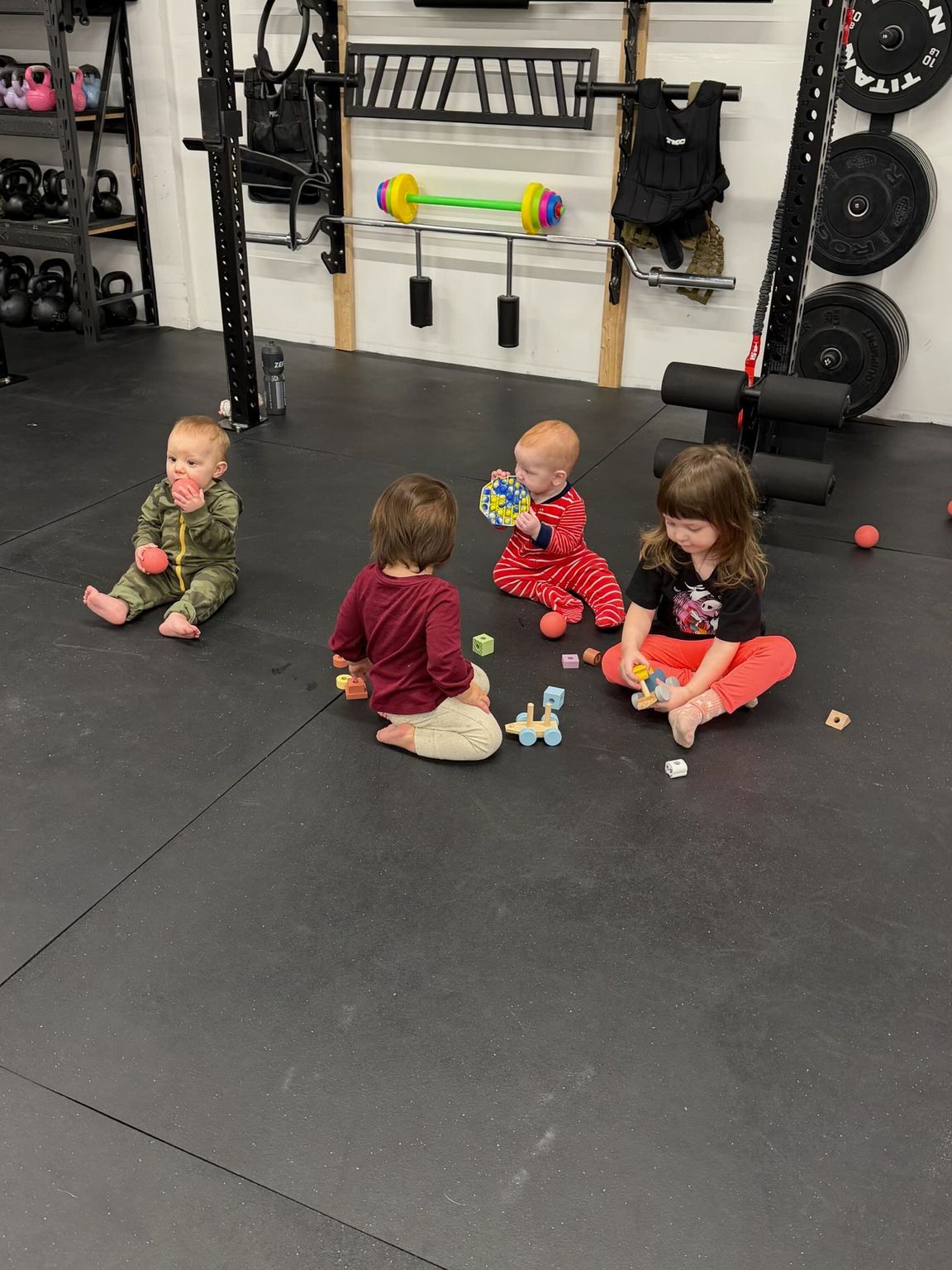 Four children sitting on a gym floor with toys, facing away from the camera, in a fitness room in Everyday Strength, a gym in Wenatchee, WA
