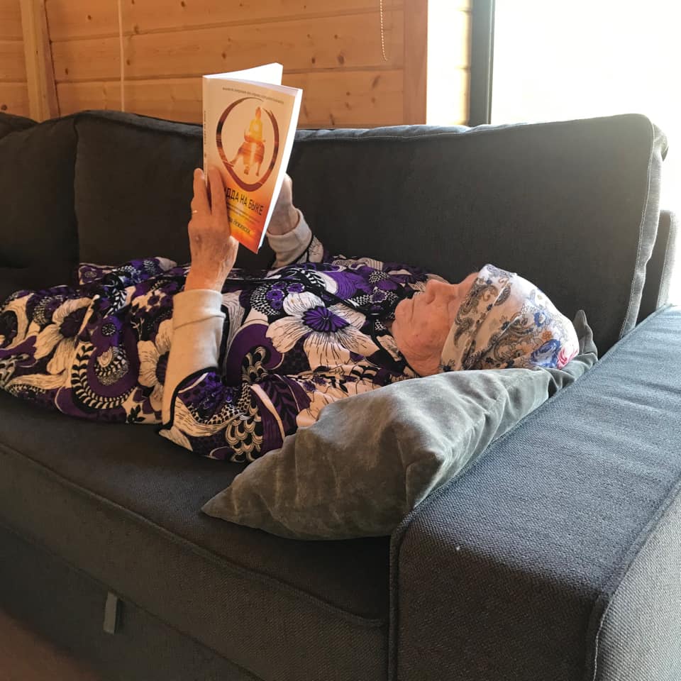 Person reclining on a gray couch, reading a book with an orange cover; sunny interior.