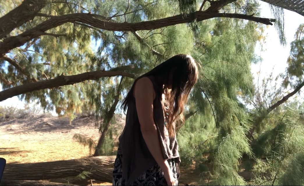 Person in a black dress standing under tree branches in a sunlit outdoor setting