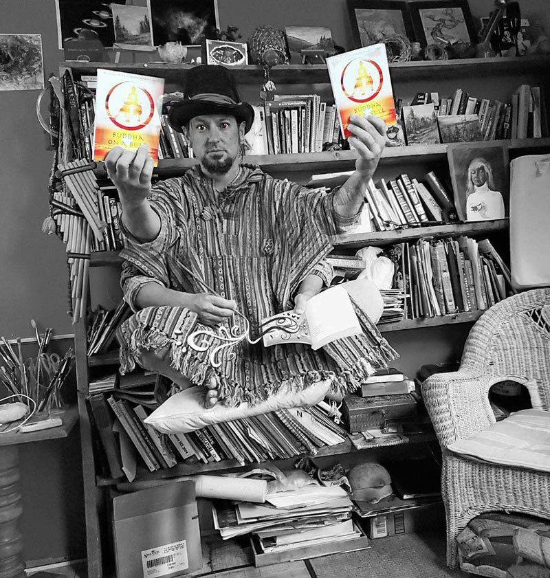 Man levitating, holding two book covers, in front of a bookshelf filled with books and art supplies.