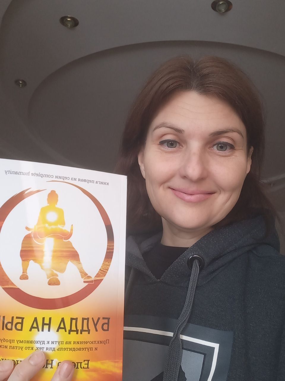 Woman holding a book with a Buddhist-themed cover, smiling at the camera, indoors.