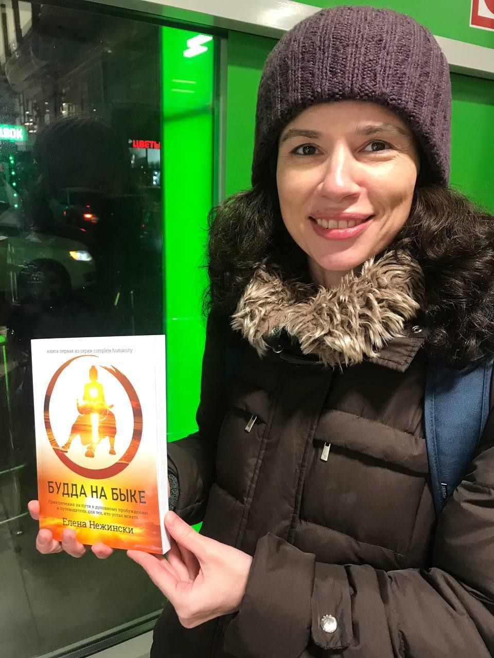 Woman in winter coat and hat holding a book with text; green backdrop.