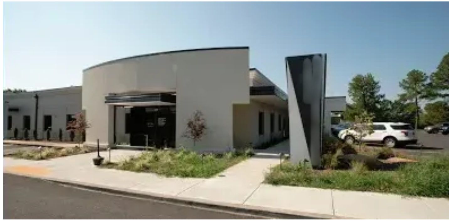 Modern building entrance with white facade, black accents, landscaped walkway, and parked cars nearby
