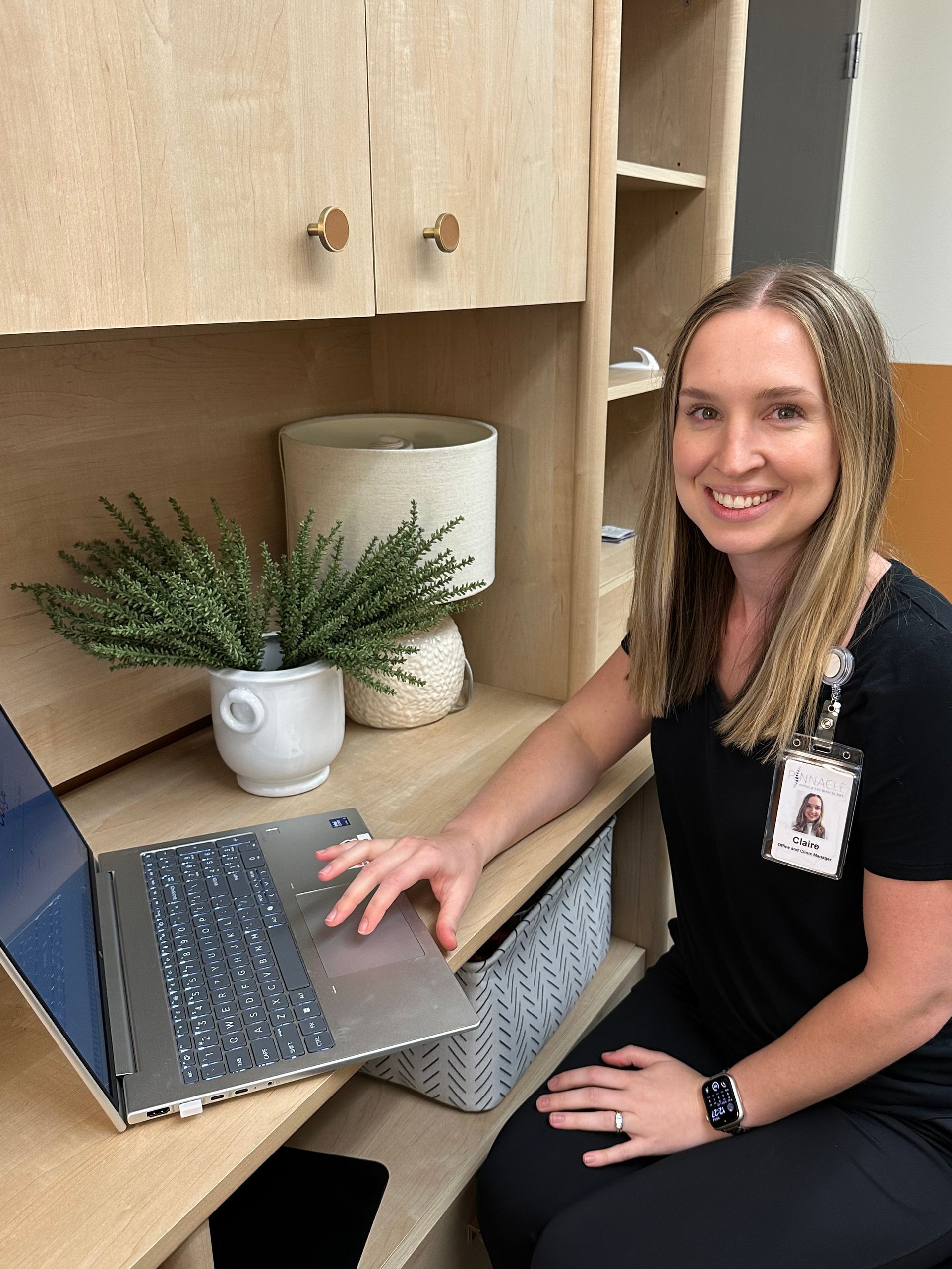 Woman smiling at a desk with a laptop, in an office setting with shelves and a plant.