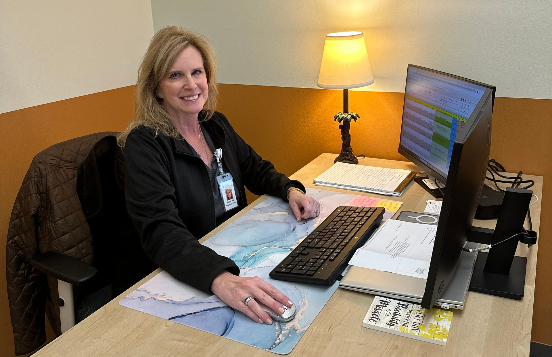 Smiling woman at a desk using a computer in an office, with a lamp and paperwork nearby