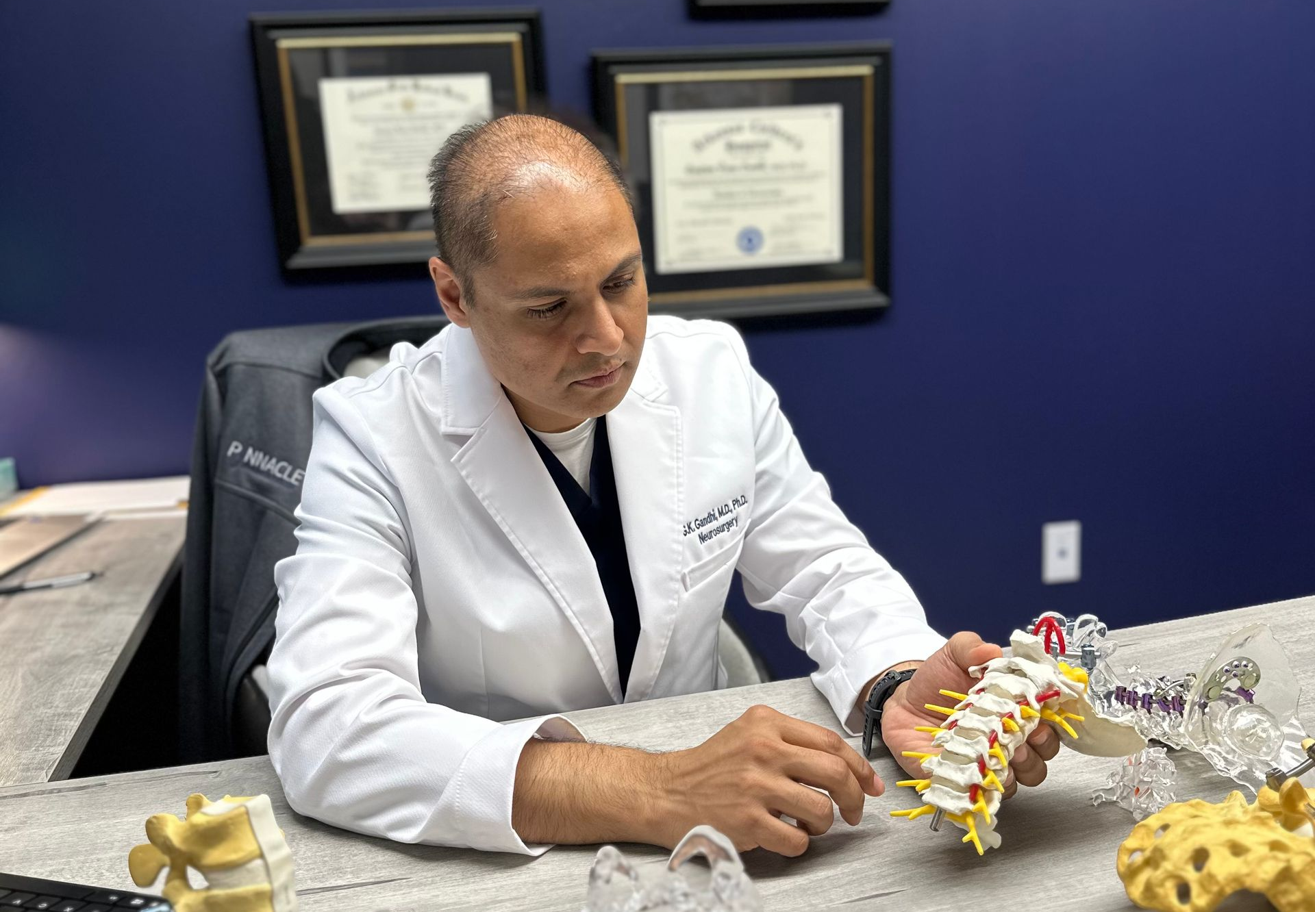 Doctor in white coat examining a spinal model at a desk in an office with framed certificates.