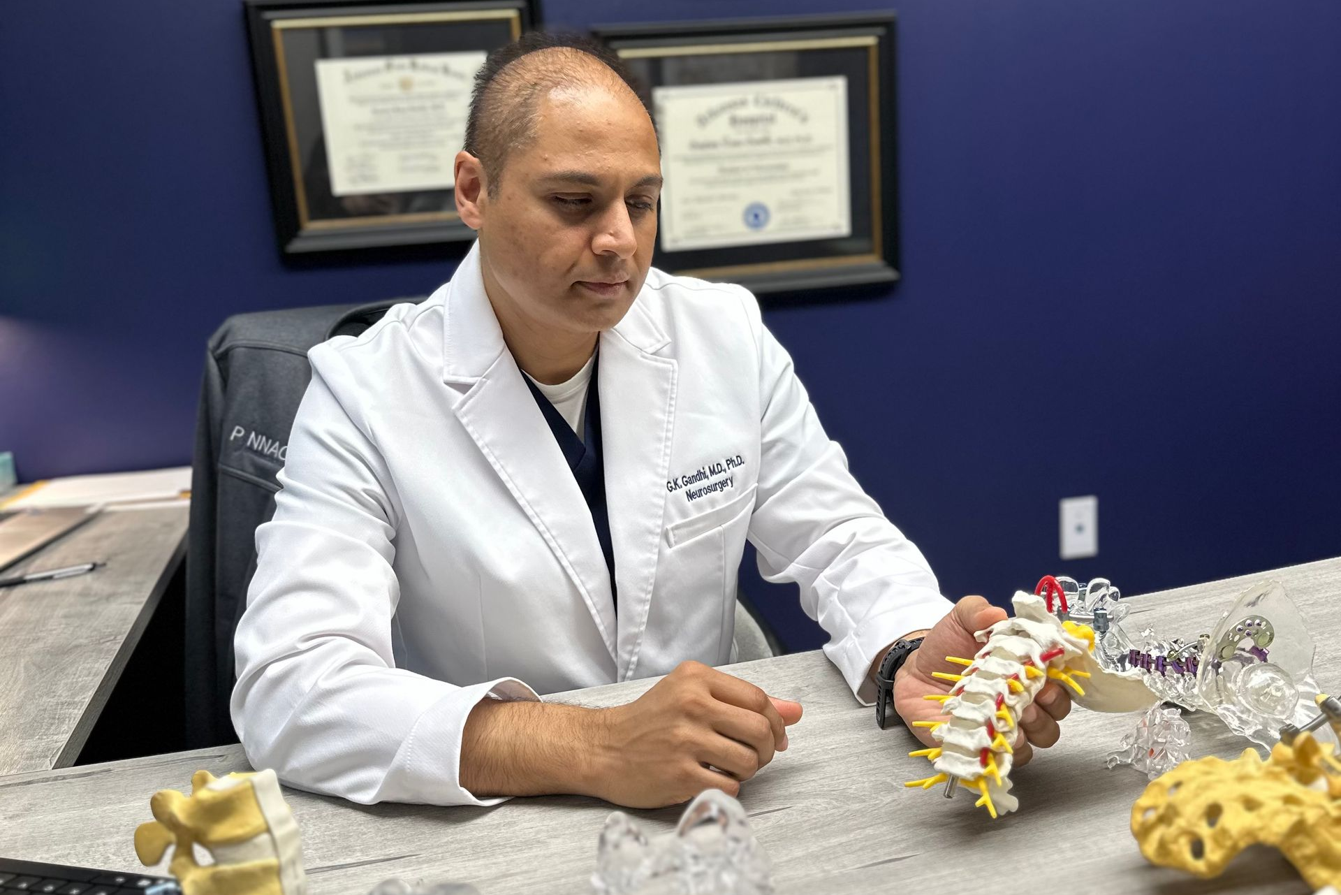 Dr. Gandhi in a lab coat, seated in his office, holding a spine model.