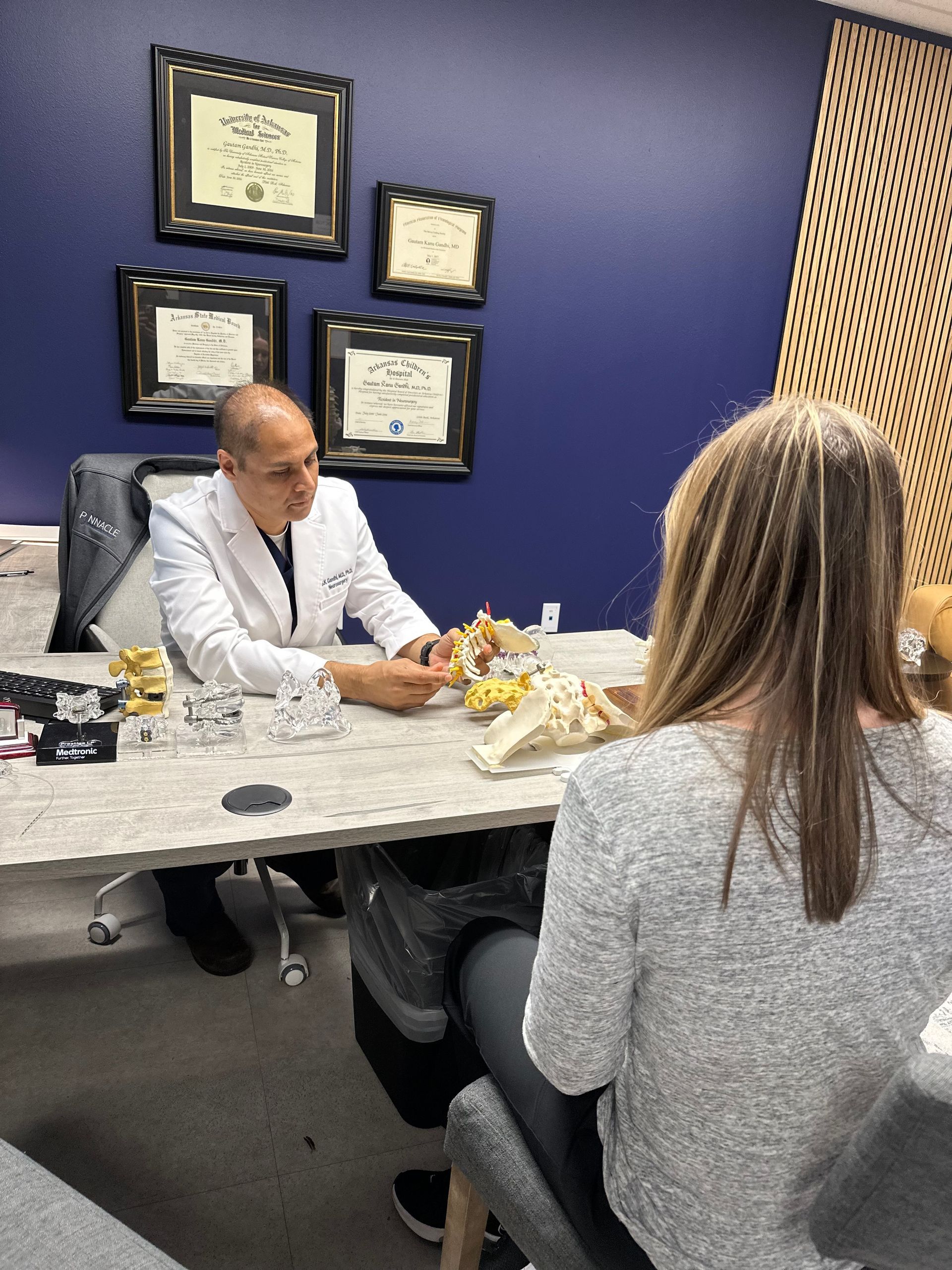 Two people meeting in a doctor's office with framed certificates on a blue wall and spine models on the table
