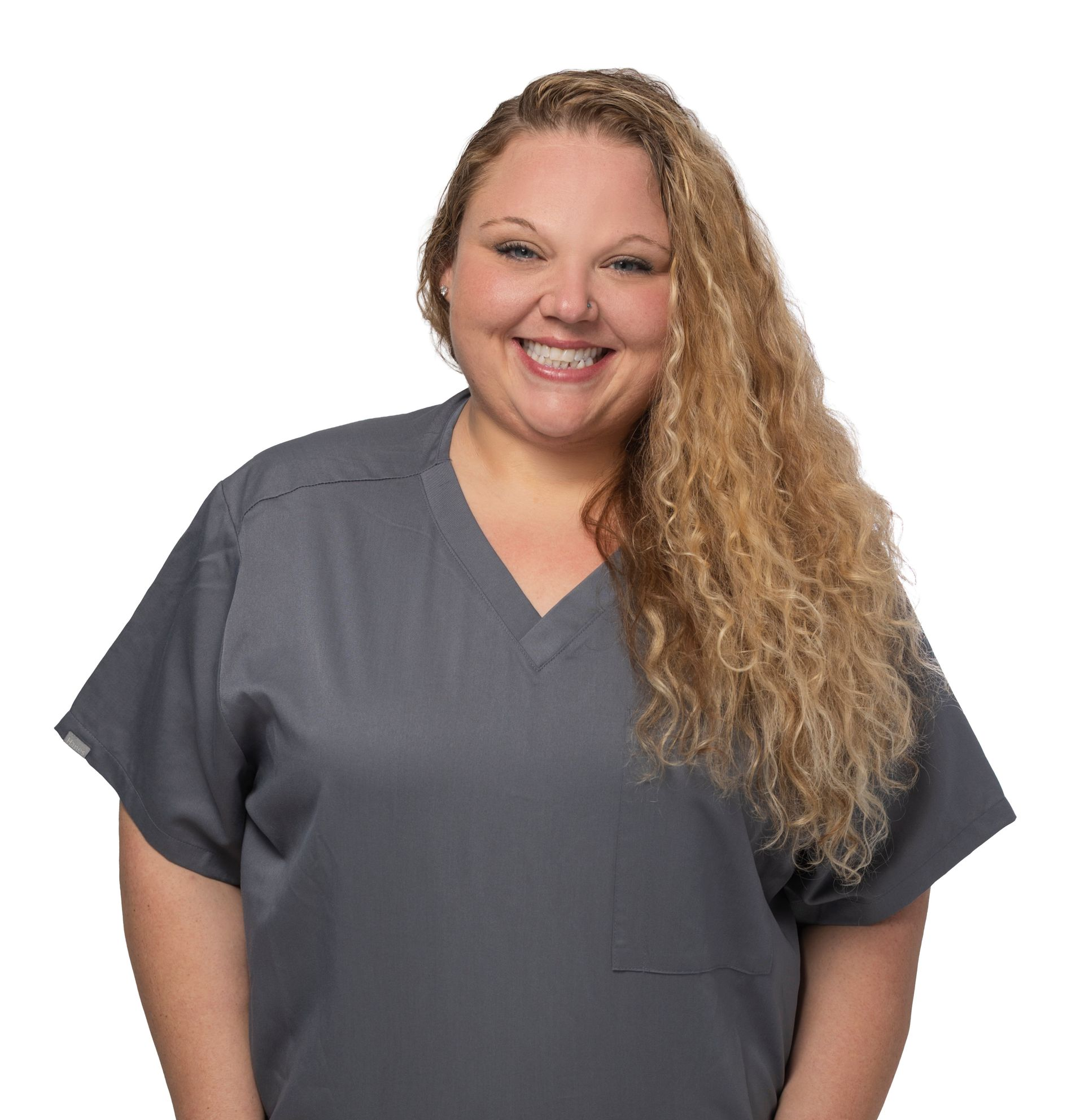 Smiling person in gray scrubs with hands in pockets against a white background.