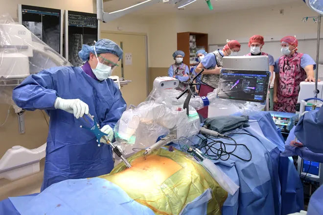 Medical team in blue scrubs performs surgery in an operating room, with monitors and surgical lights visible