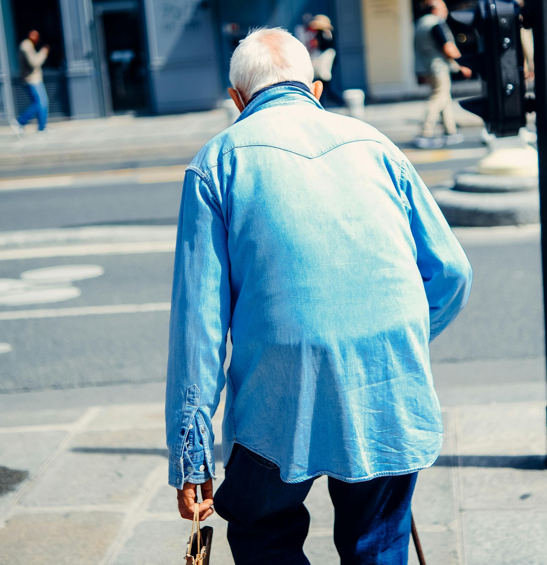 Person in a light blue shirt walking away on a city sidewalk, holding a cane.