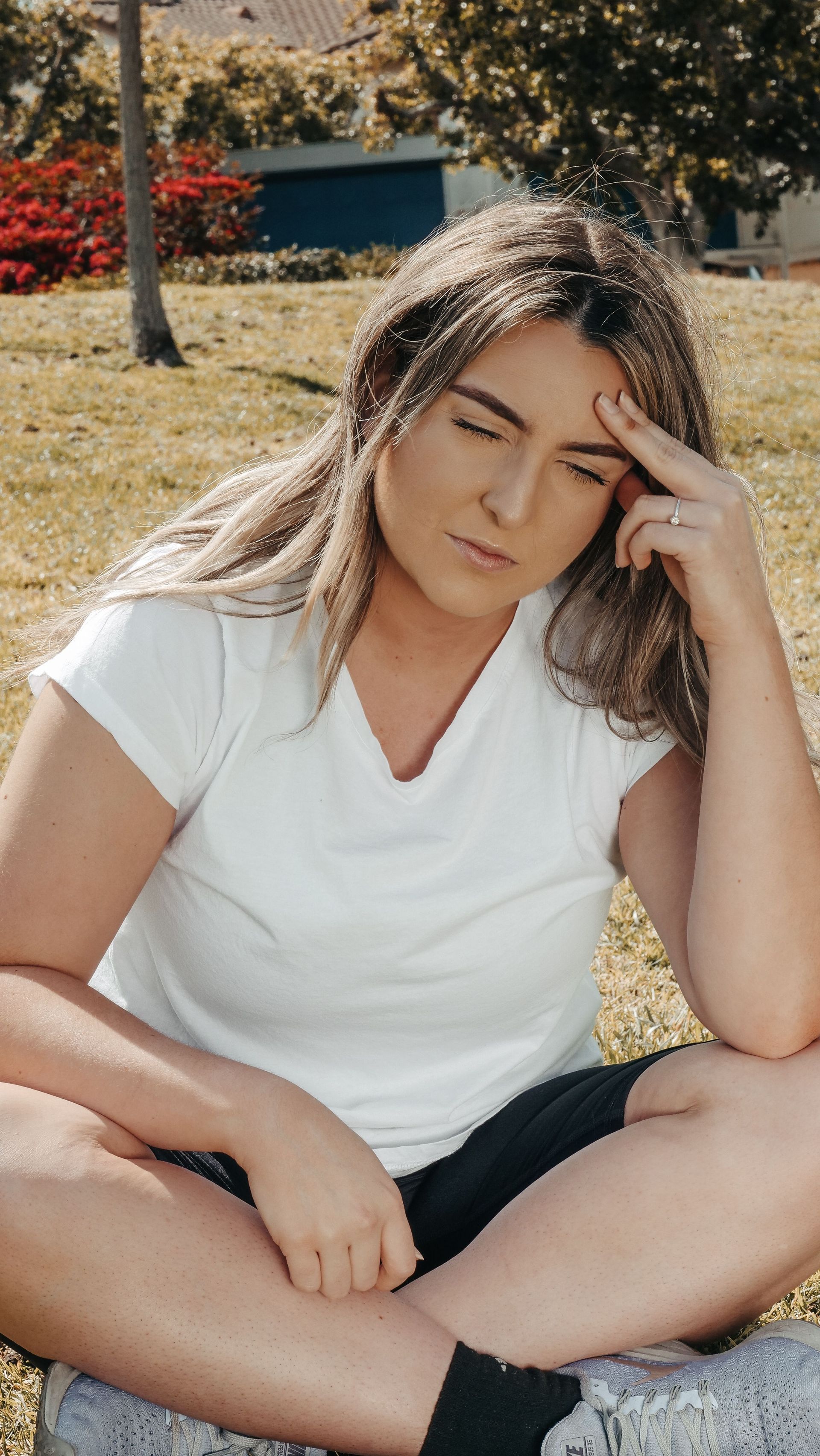 Woman sitting cross-legged on grass, looking down and touching her forehead, wearing a white shirt and black shorts