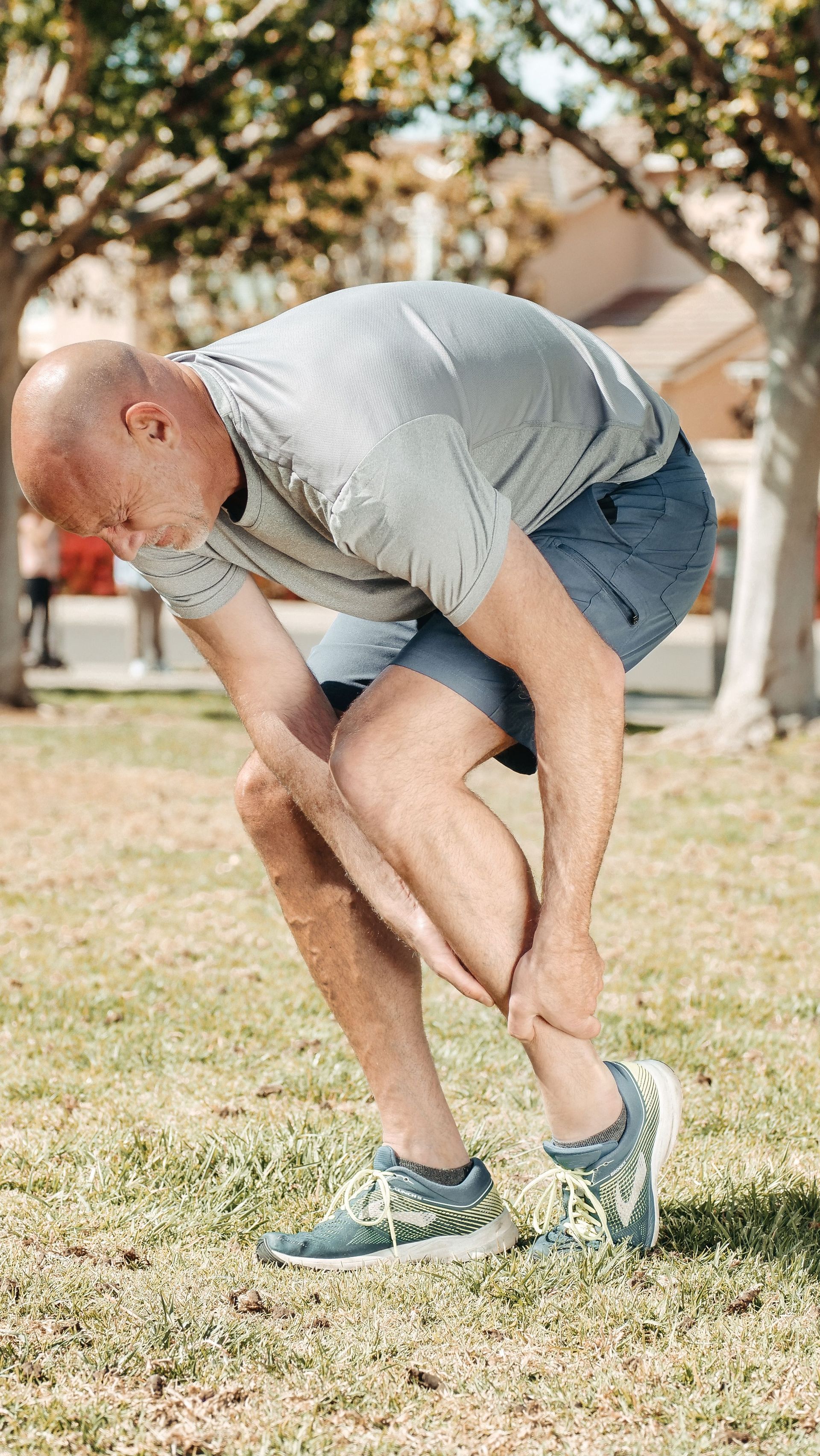 Man in gray shirt and blue shorts bending over, holding his leg on a grassy field.
