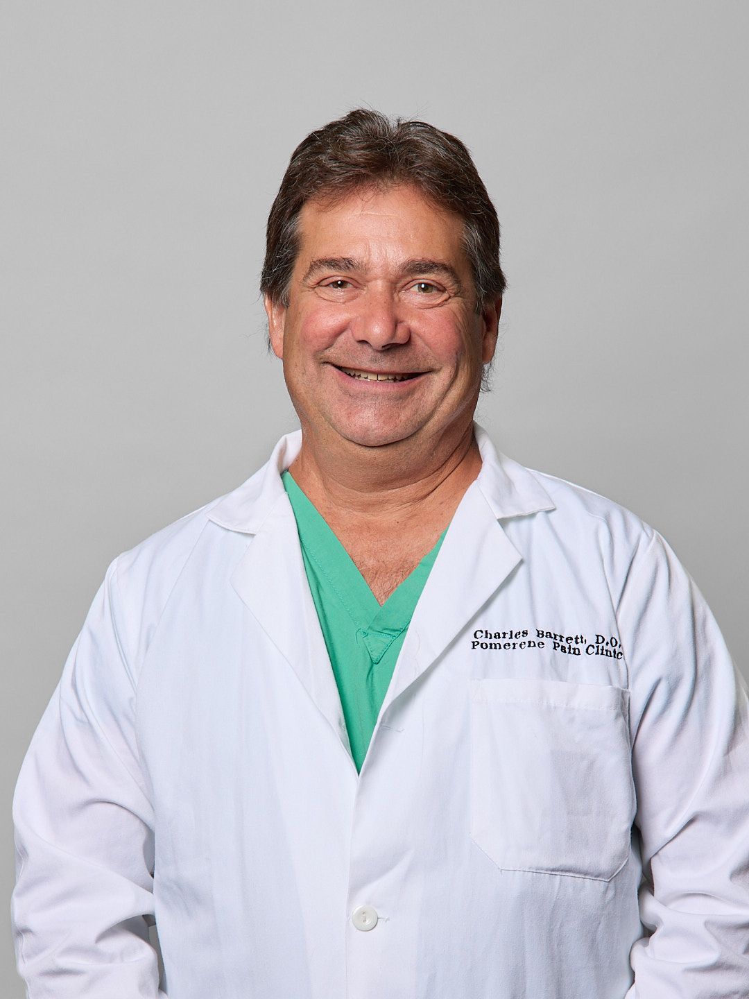 A smiling doctor in a white coat and green scrubs stands in front of a gray backdrop.