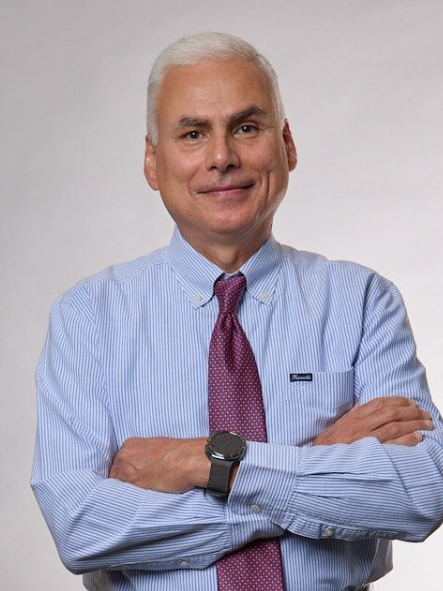 A middle-aged man with gray hair, wearing a blue striped shirt and red tie, smiles at the camera.