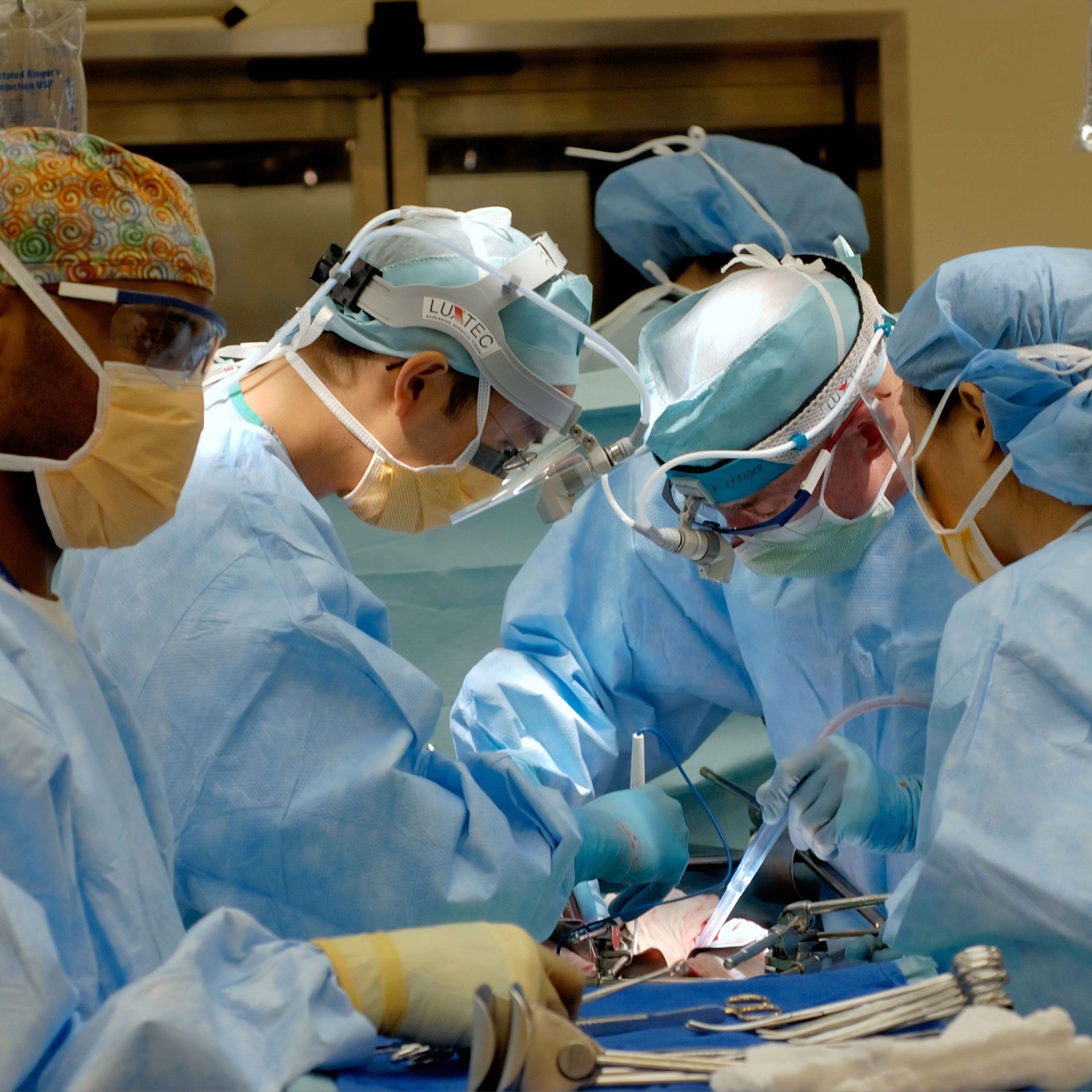 Surgeons in blue scrubs and masks operate in a bright surgical room.