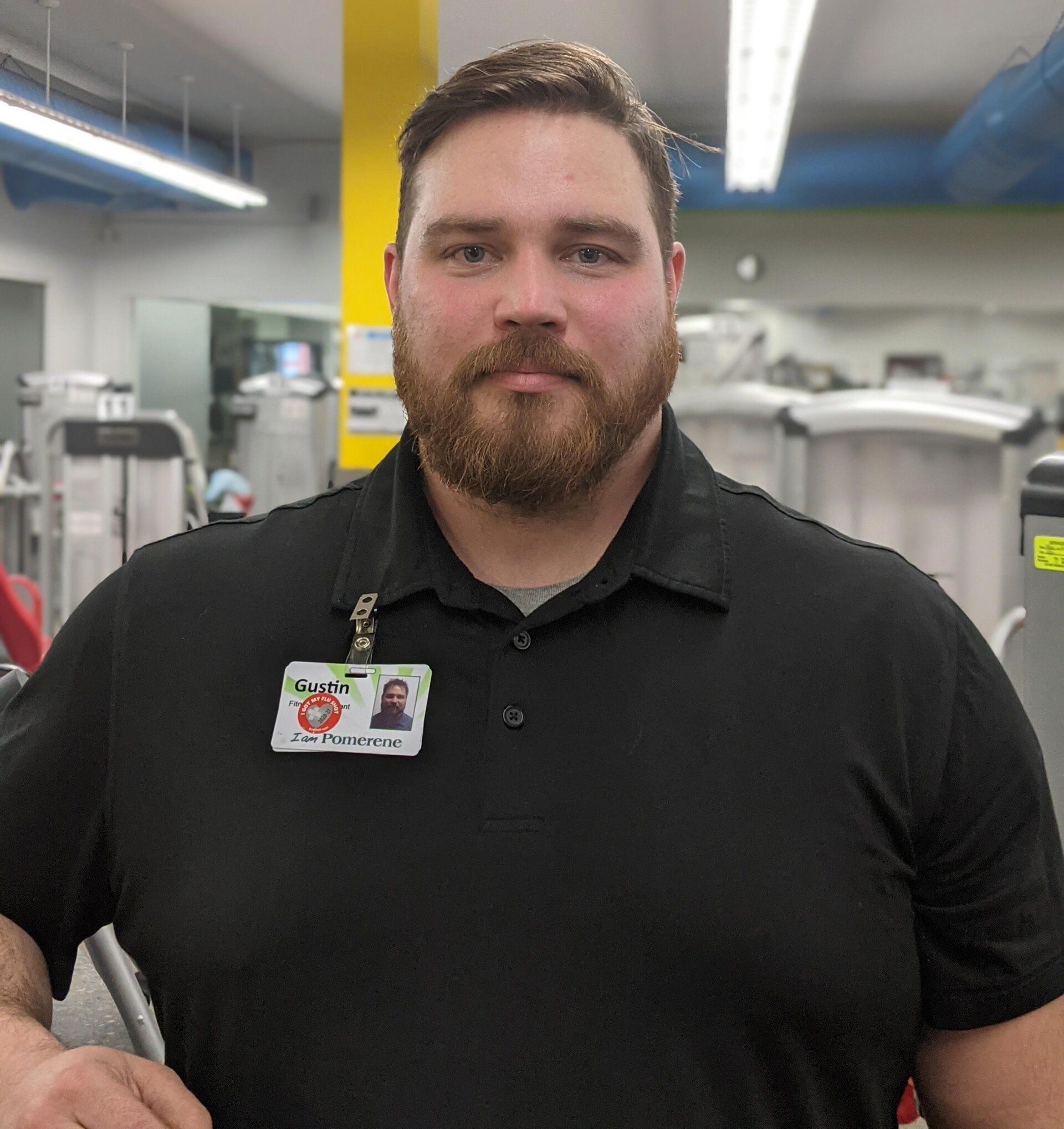 Man with beard wearing a black polo shirt and name tag, standing in a gym.