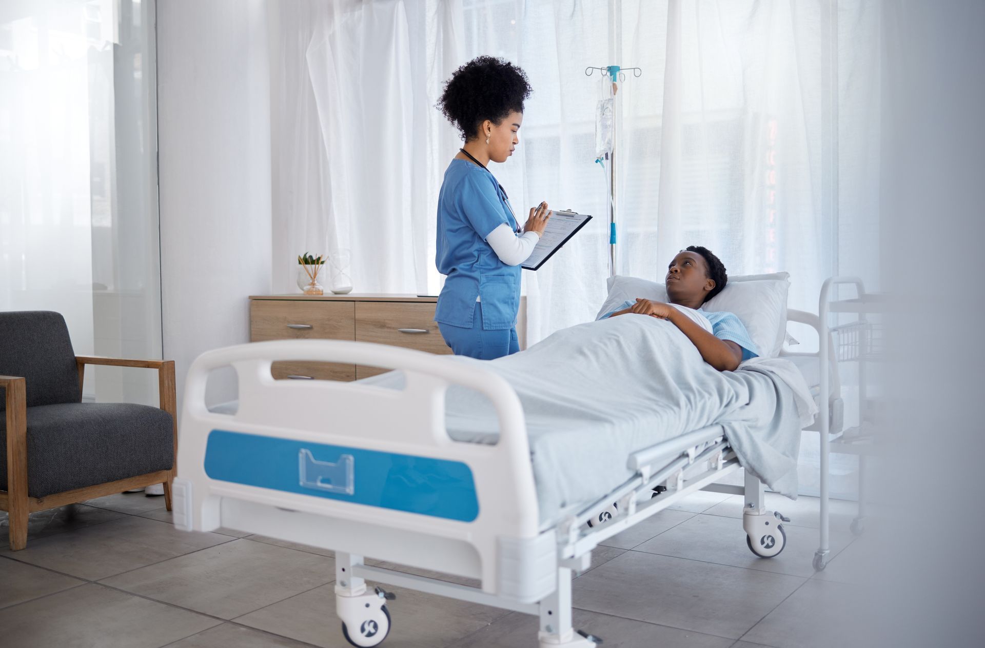 A Black nurse in blue scrubs consults with a patient lying in a hospital bed. The room is bright with natural light.
