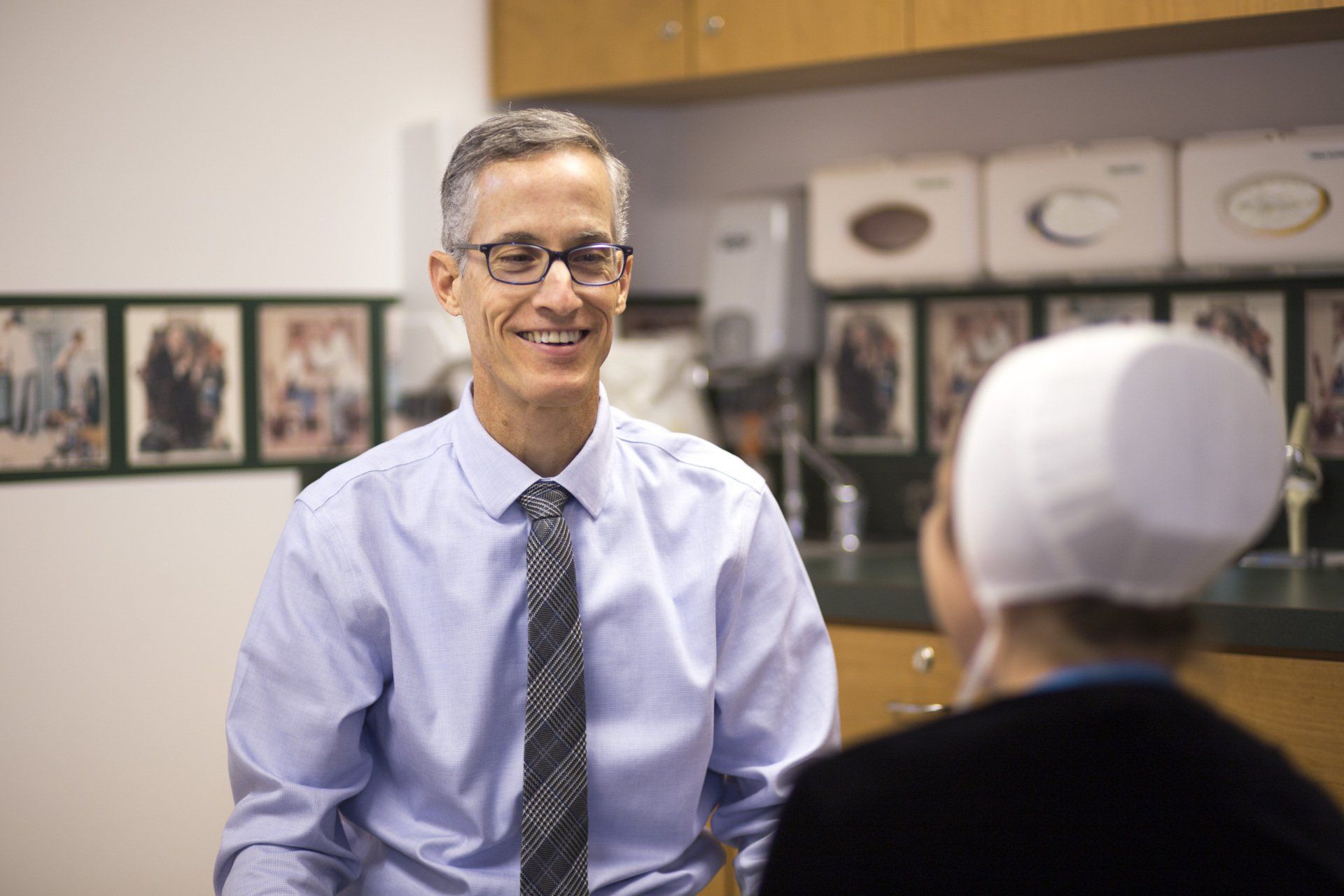A doctor in glasses smiles at a patient wearing a white head covering in an office setting.