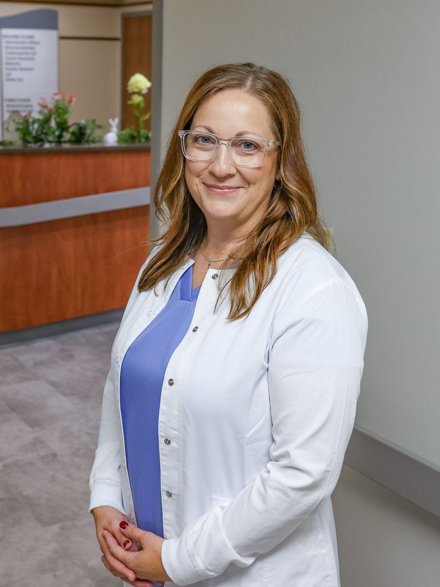 Woman with glasses, wearing a white lab coat over a blue top, smiling in a hospital hallway.