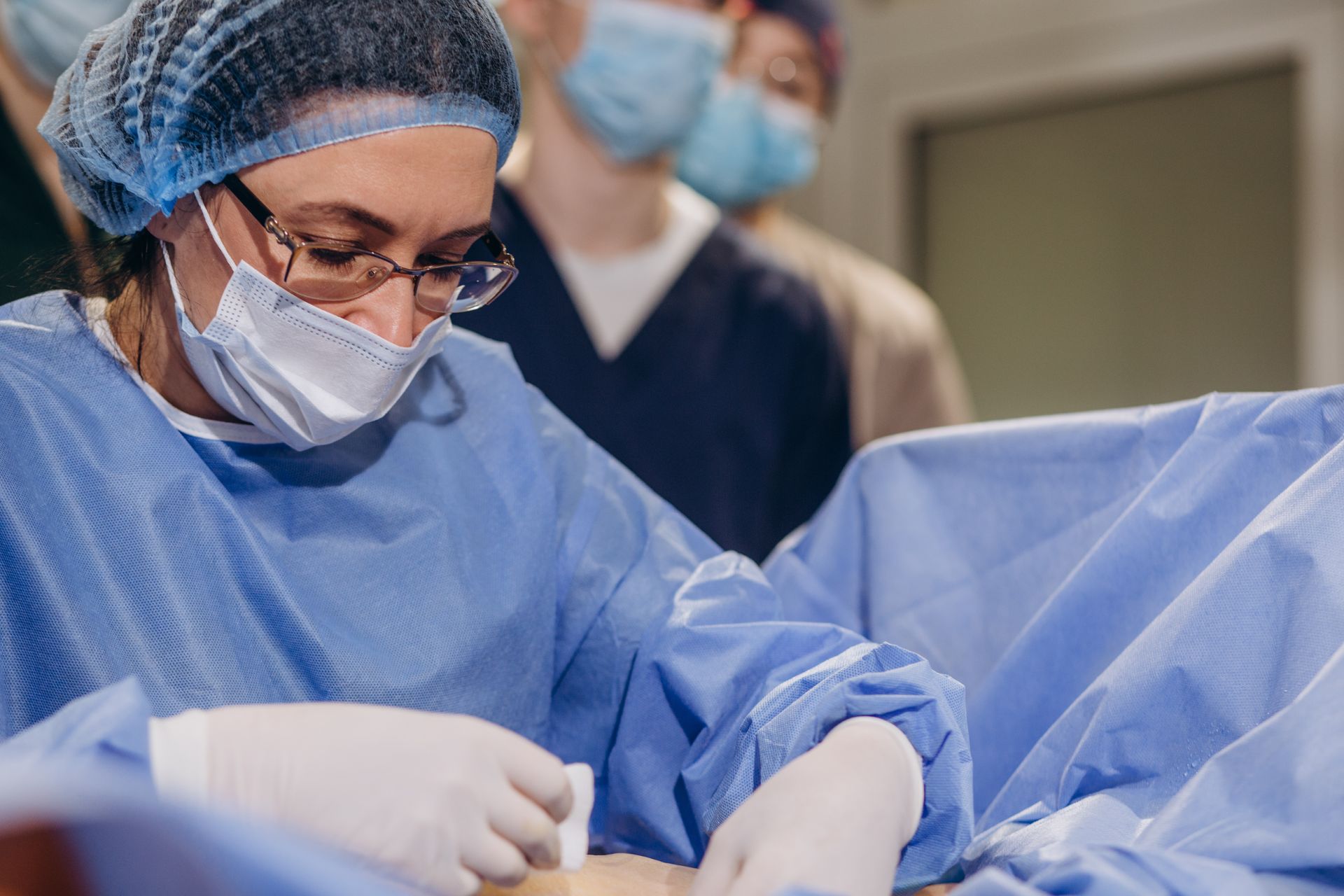 Surgeon in blue scrubs, hairnet, and mask, performing a procedure. Other medical staff are in the background.