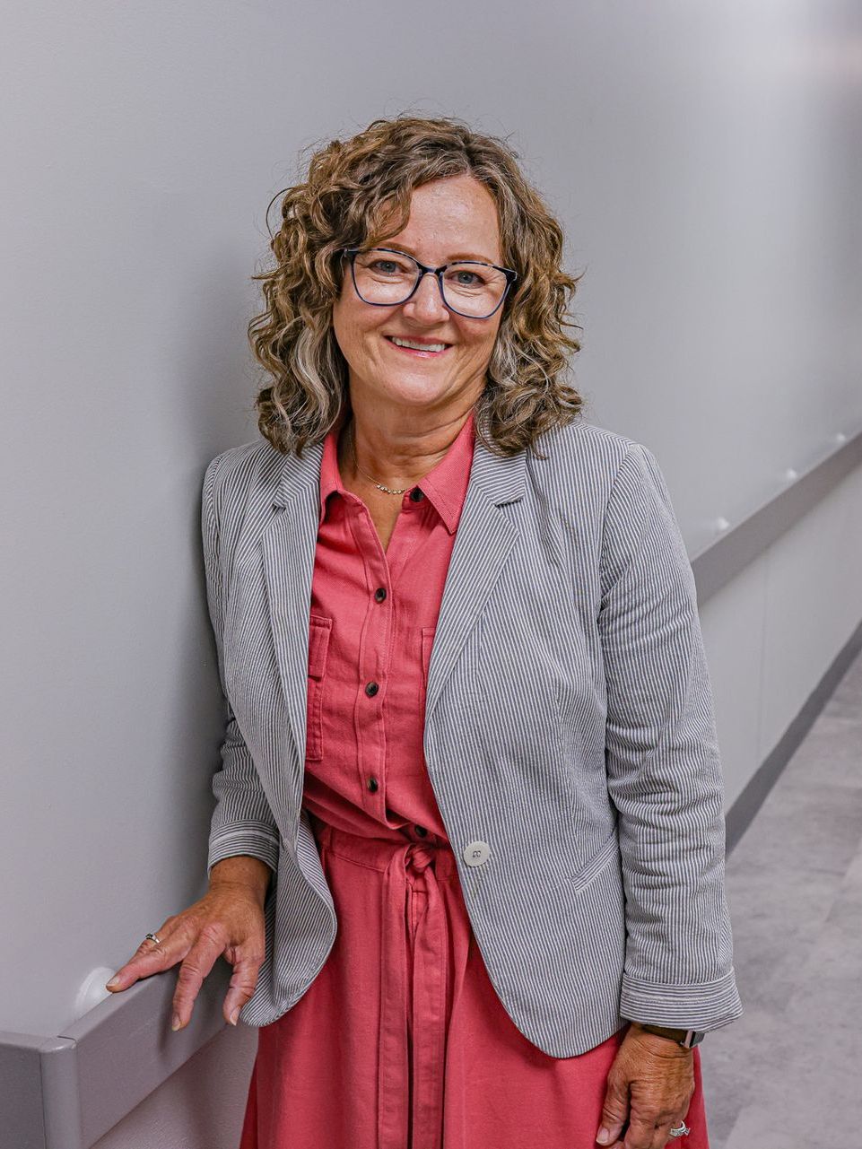 Woman with curly brown hair wearing glasses, a striped blazer, and a pink dress, smiles while leaning against a wall.