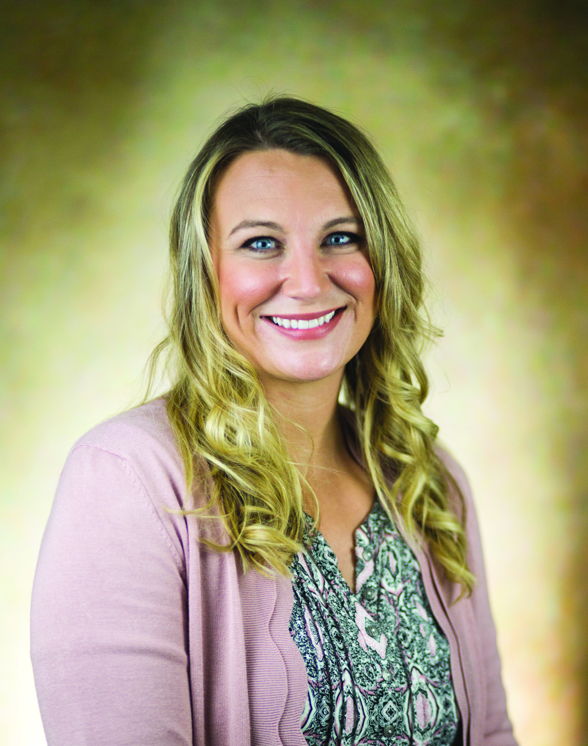 Woman with blonde hair, smiling, wearing a pink cardigan and patterned shirt, set against a blurred, golden background.