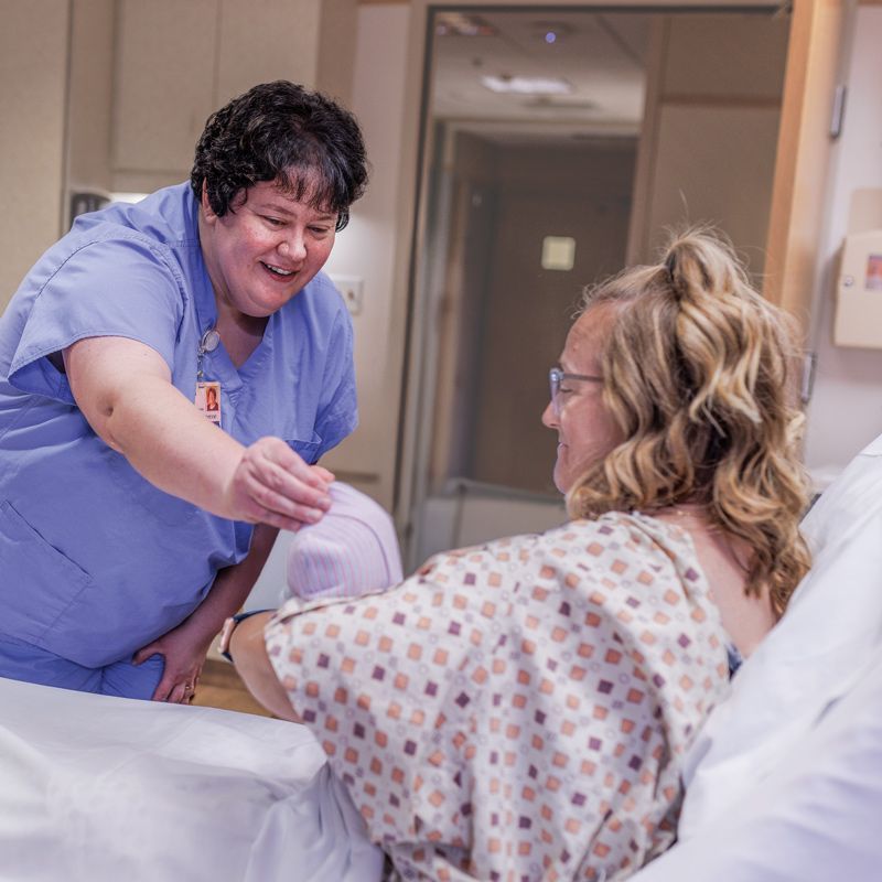 Nurse in blue scrubs smiles at a woman in a hospital bed, handing her a baby swaddled in pink.