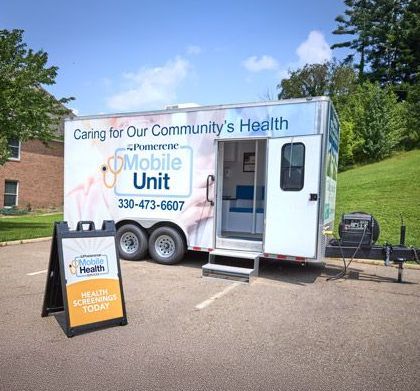 Mobile health unit trailer with open door and sign for health screenings. Located outdoors in a parking lot.