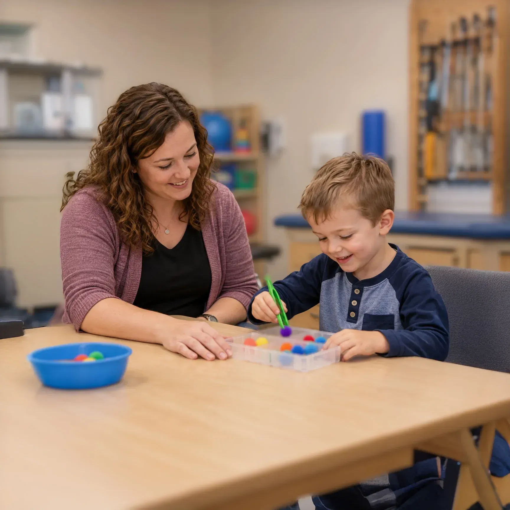 Woman in green shirt and young girl smiling, playing with clay on a wooden table in a room with a computer and printer.