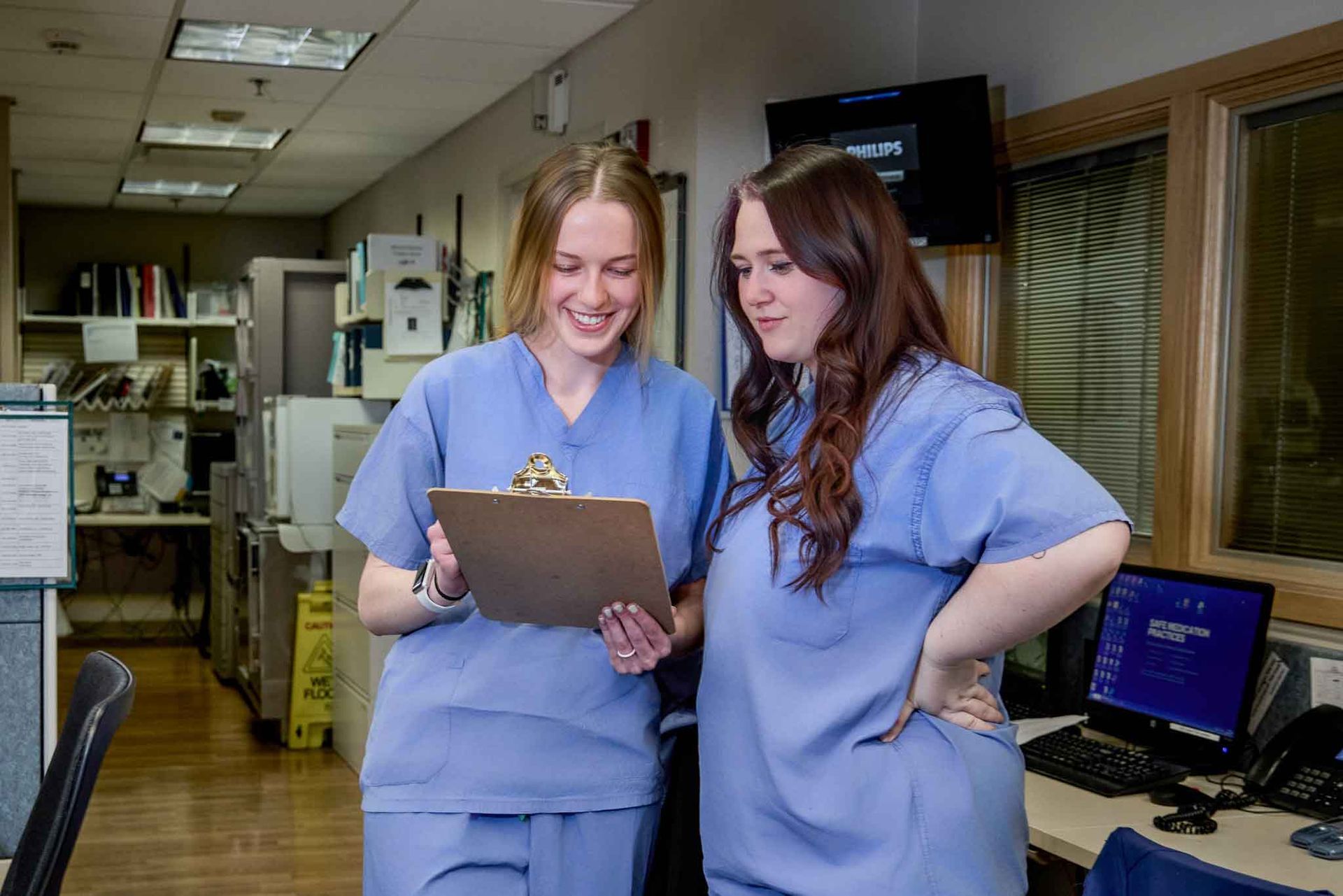 Two women in blue scrubs reviewing a clipboard, smiling in a medical office setting.