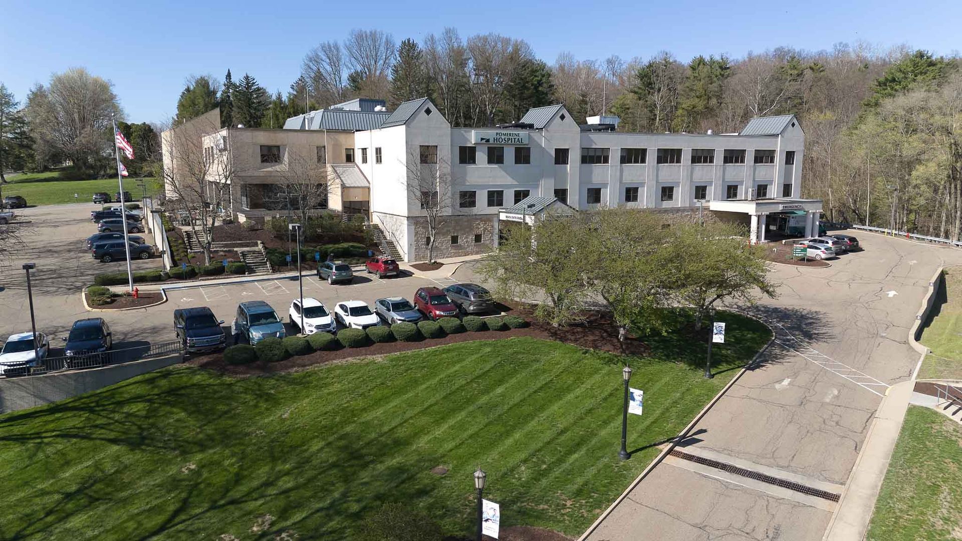 Exterior view of hospital building with multiple parked cars in front, a lawn, and trees in the background.