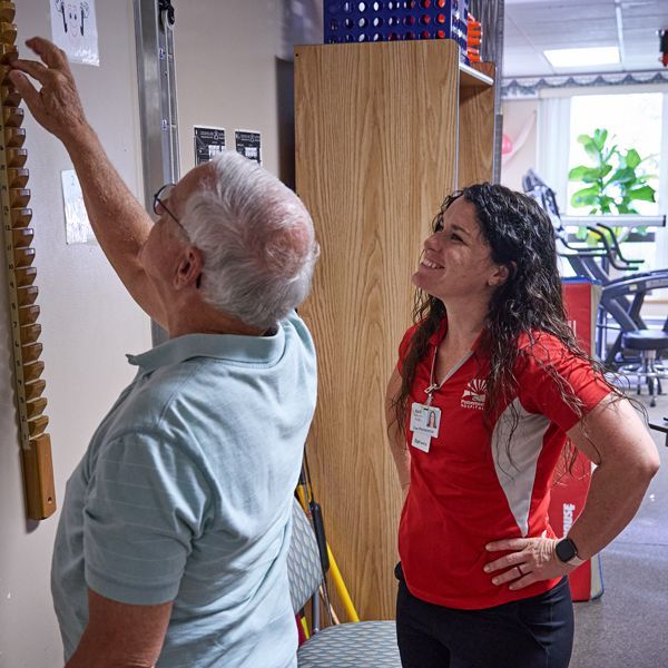 An older man reaches toward a wall-mounted exercise device while a woman watches with a smile in a physical therapy room.
