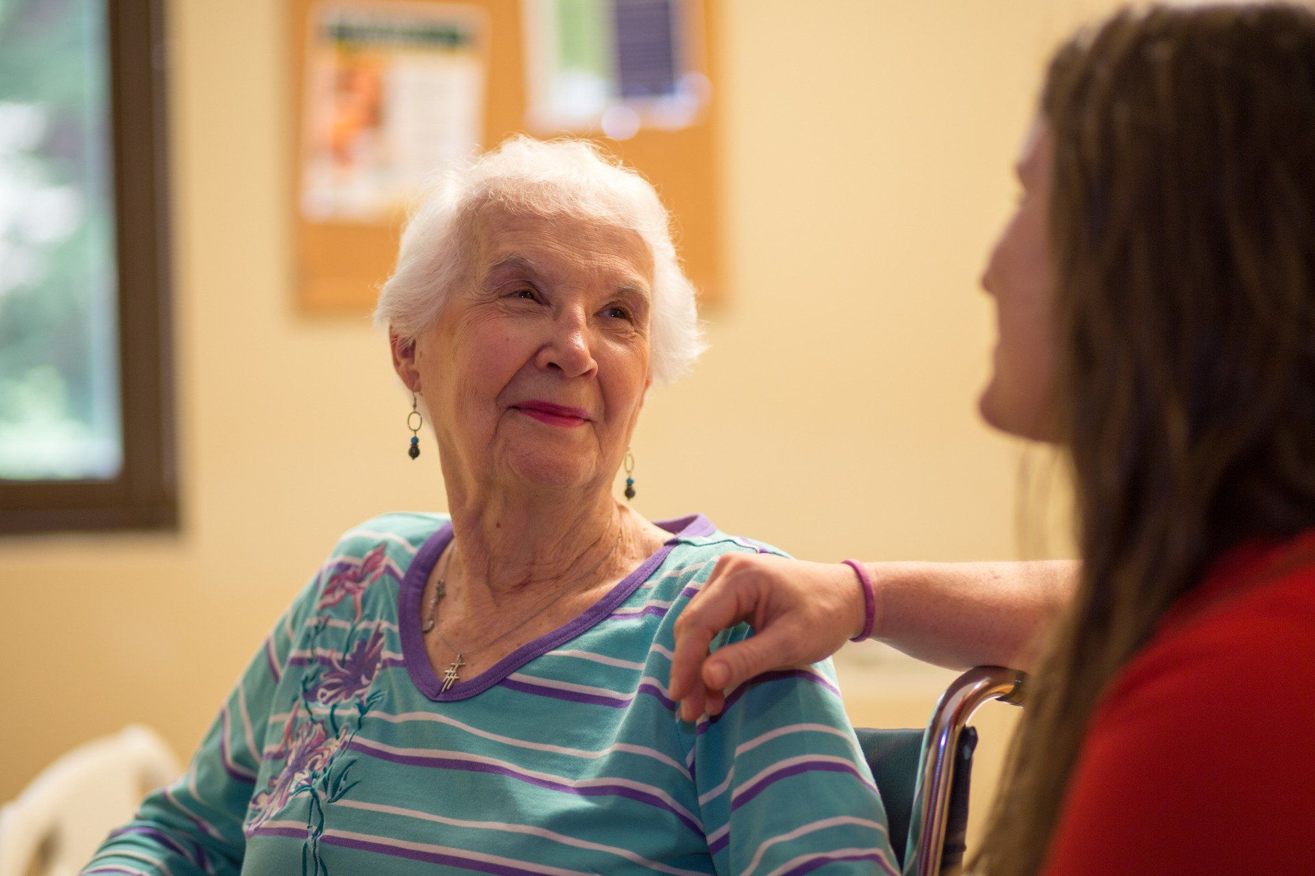 An older woman smiles as a younger woman rests her hand on the older woman's shoulder. Indoors, in a room.