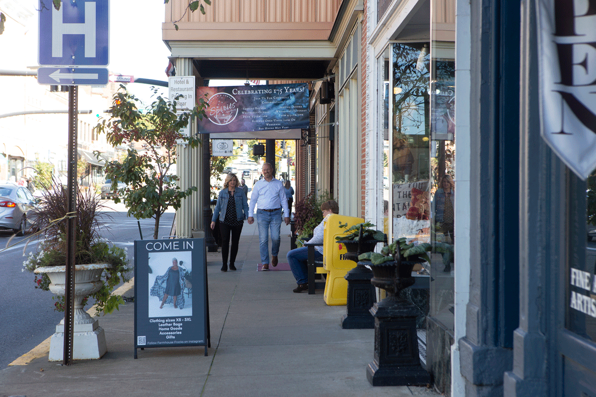 A sidewalk with shops, a couple walking, and a man sitting. A sign for a hospital is visible.