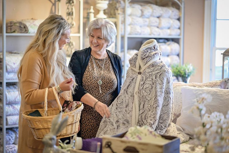 Two women shop at a store. One holds a basket, while the other examines a lace shawl.
