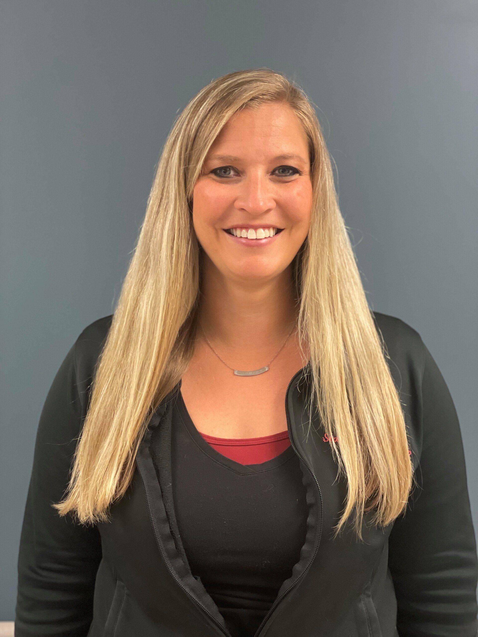 Blonde woman smiling, wearing a black jacket and maroon shirt, in front of a grey wall.