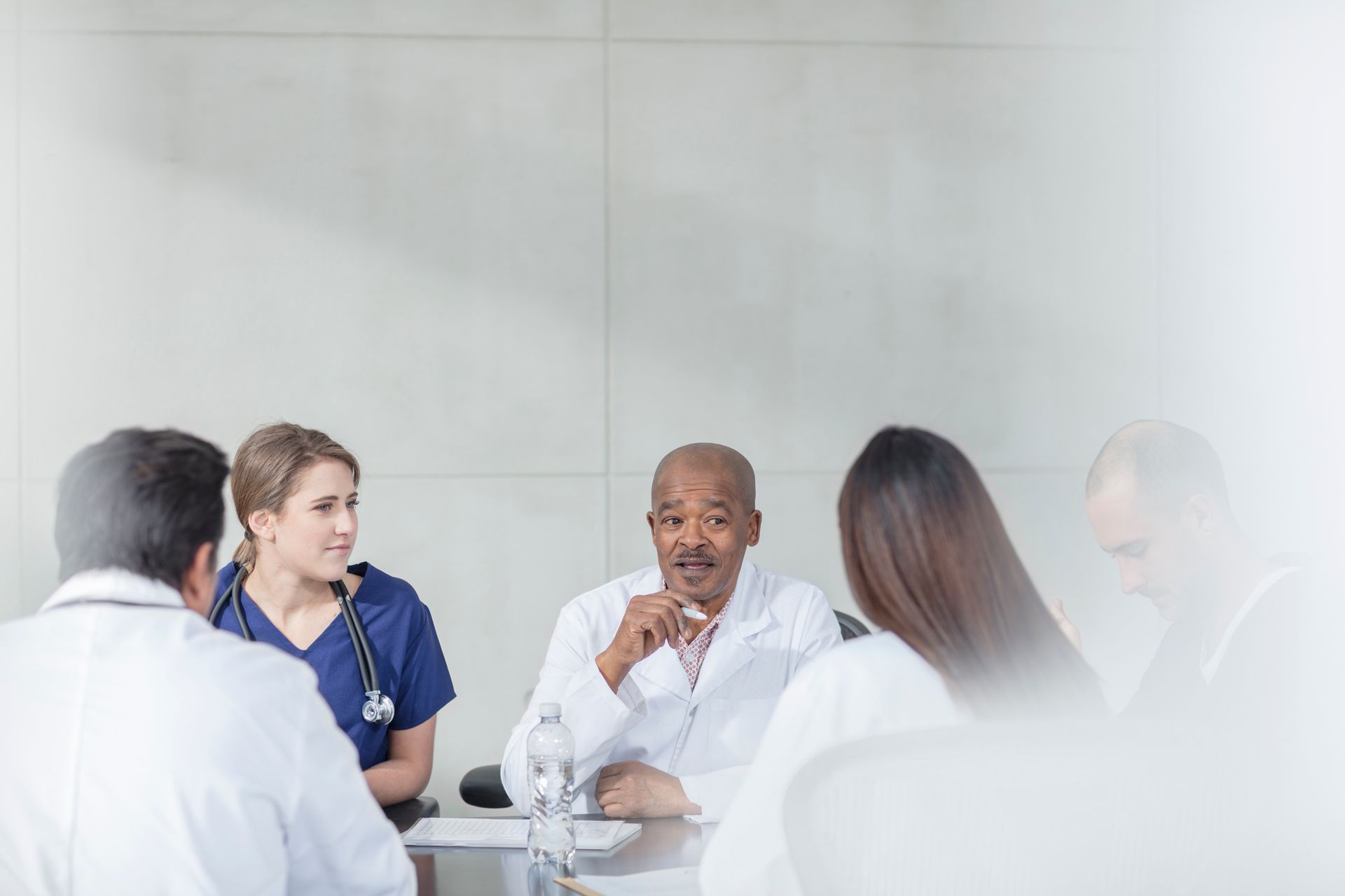 Medical professionals in white coats and scrubs sit at a table, engaged in a discussion. Light background.