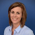 Woman with short brown hair smiling at the camera, wearing a blue button-down shirt, with a blue background.
