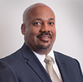A Black man in a suit and tie smiles at the camera, chest up, against a white background.