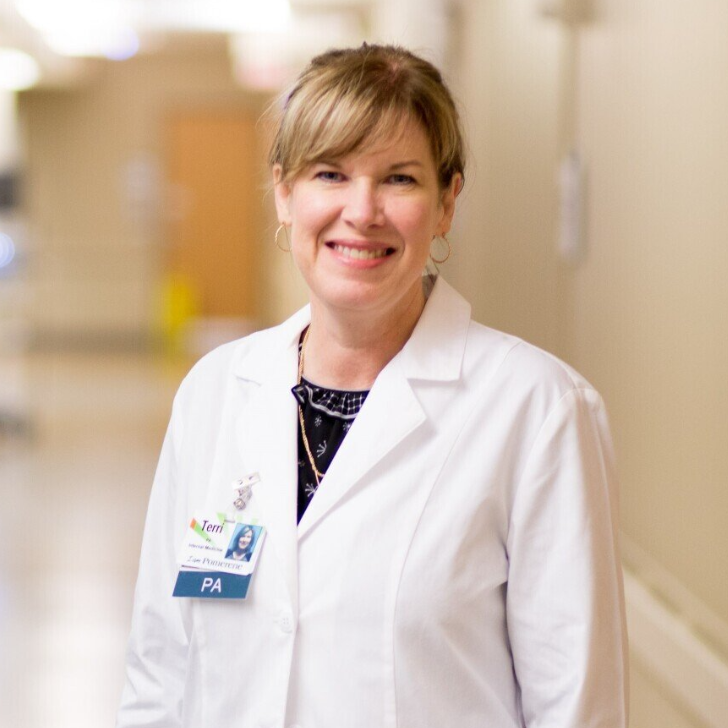A woman in a white coat, a PA badge, smiles in a hospital hallway. She has blonde hair and wears a patterned blouse.