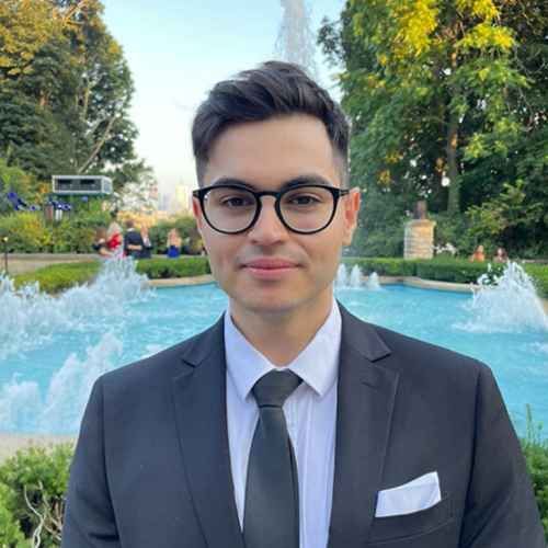 Man in a navy suit and glasses standing by a fountain in a park.
