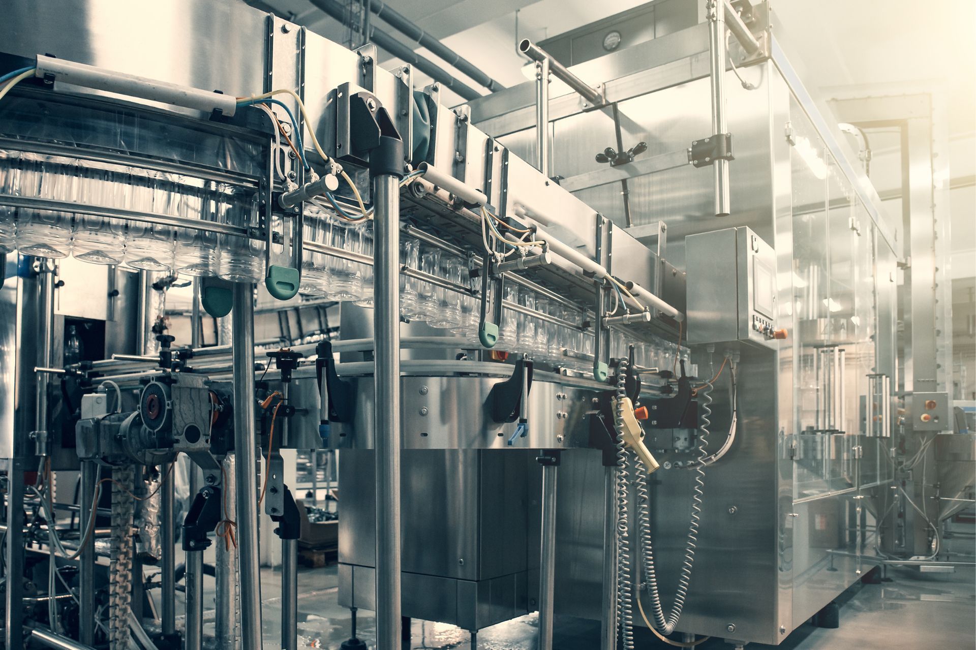Industrial factory floor with metal machinery, pipes, and overhead conveyor lines in a bright, smoky room
