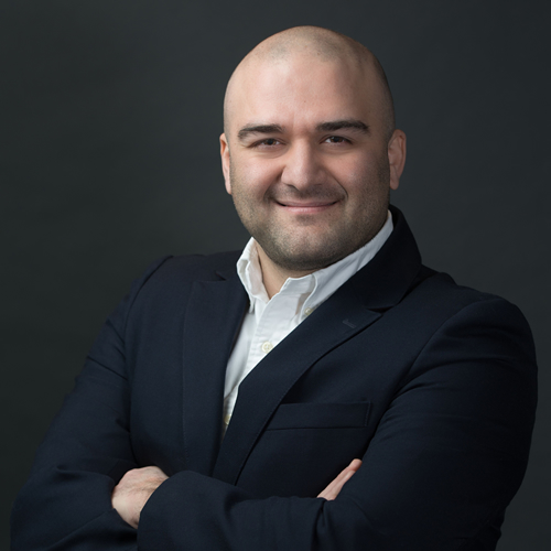 Khaled Baranbo | Manufacturing Business Broker in Ontario, Canada Smiling bald person in a dark suit with arms crossed against a dark background