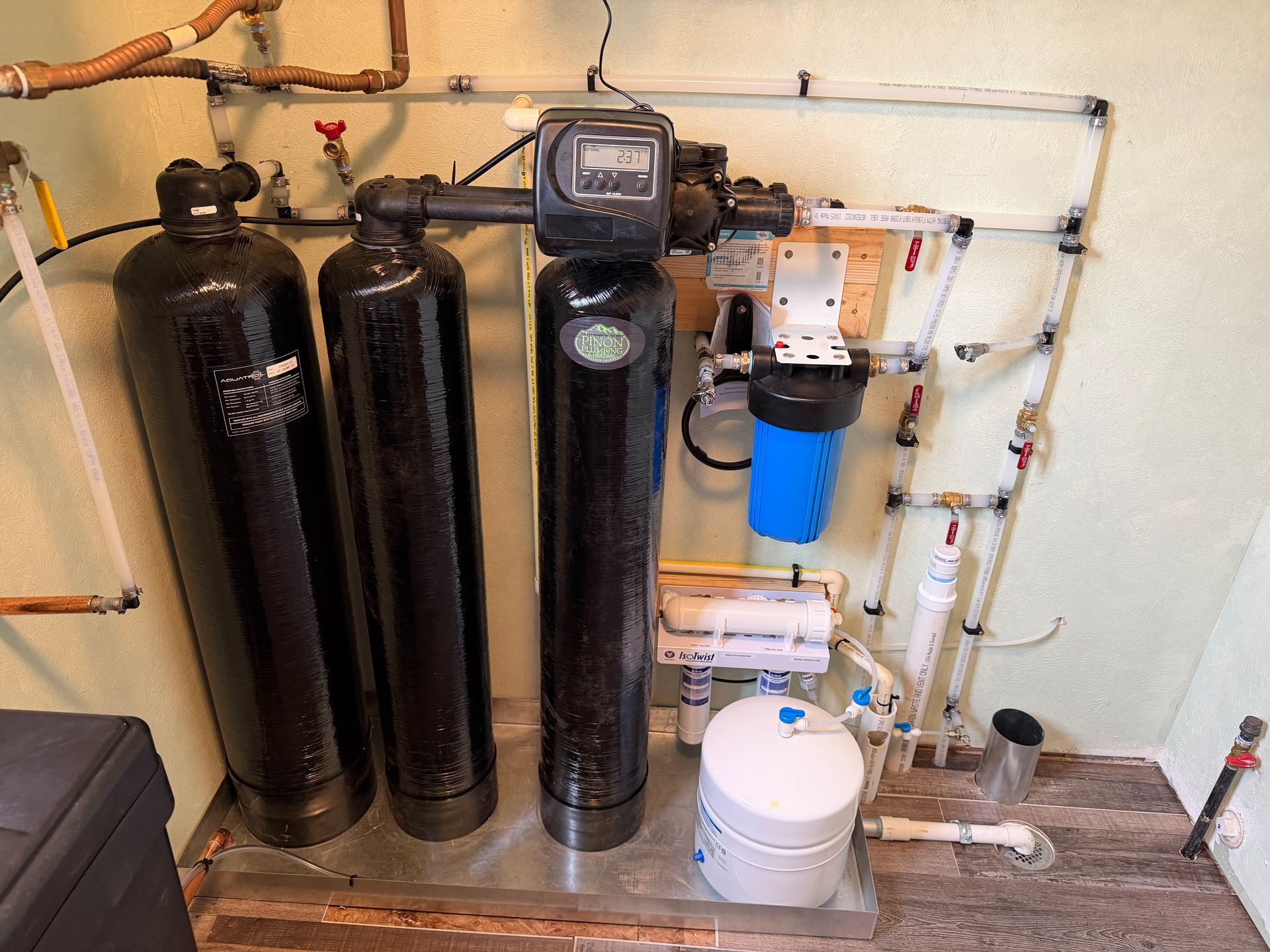 Water softener and filtration system with black tanks, blue filter, and white pump in a utility room.